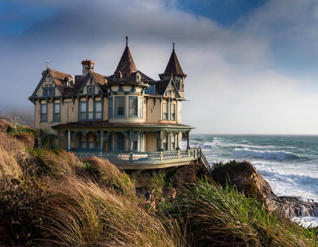 A new Victorian-style house perched precariously on a cliff's edge, overlooking an ocean with crashing waves under a partly cloudy sky.